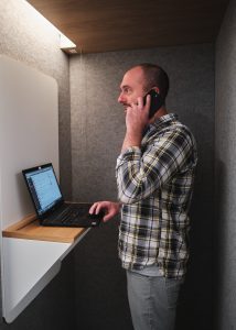 man talks on phone in a makeshift phone booth with laptop stand and laptop open