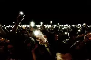 large crowd of people standing in the dark waving their cellphones with the flashlight feature enabled