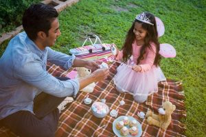 A father playing dress up with his daughter on a plaid picnic blanket.