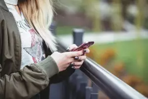 Tight shot of a women with blonde hair in a green jacket using a pink smartphone.