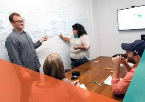 two workers standing at head of conference room writing on the wall talking to two workers sitting at a table