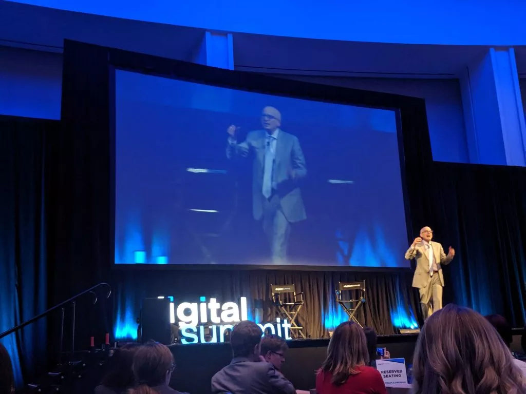 man standing on stage in front of an audience with his image projecting behind him