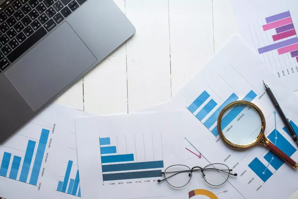 eyeglasses, computer and a magnifying rest on documents filled with charts and data to signal market research