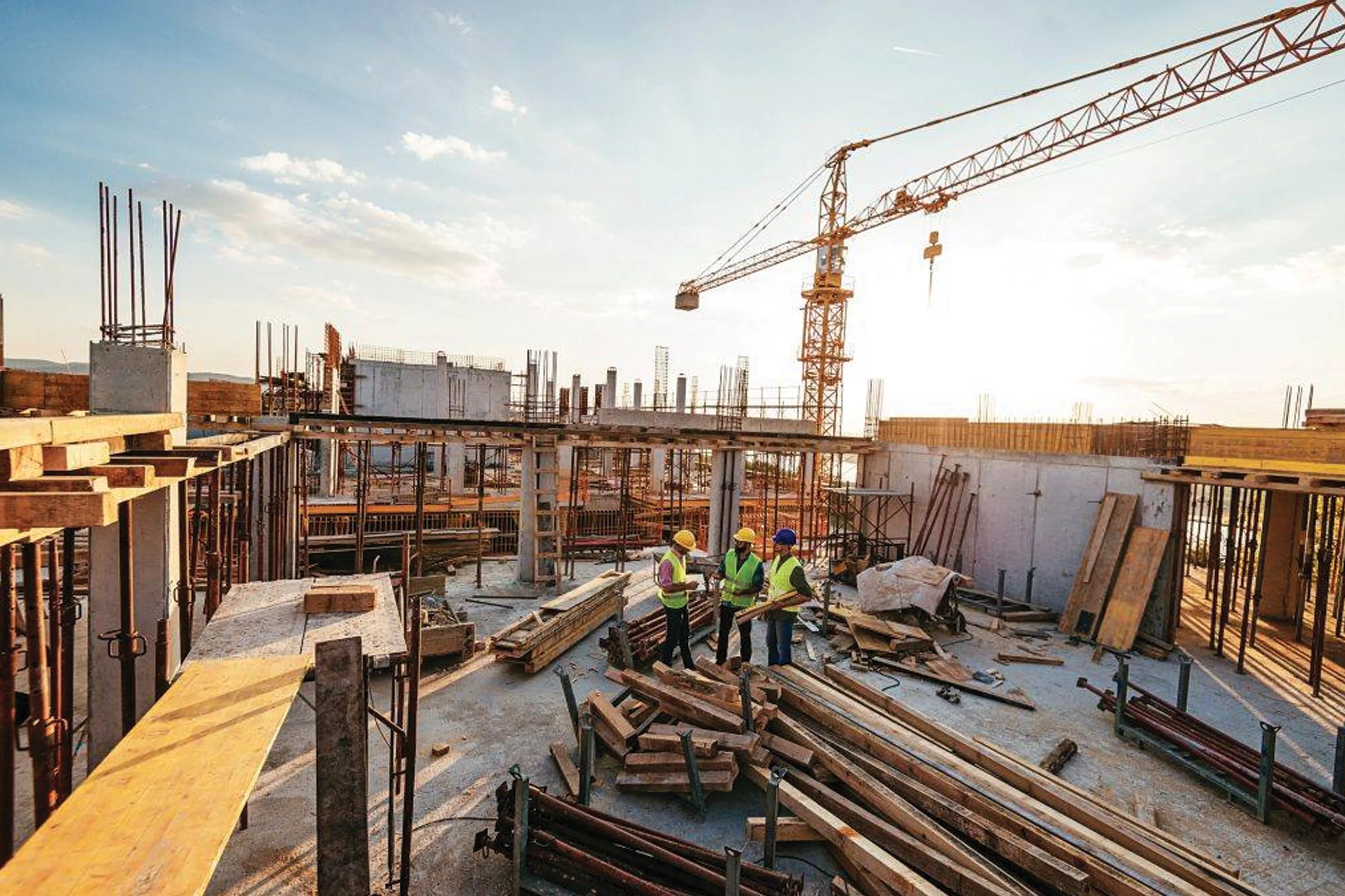 Three construction workers talk on a job site, surrounded by lumber, half-finished walls, and a crane