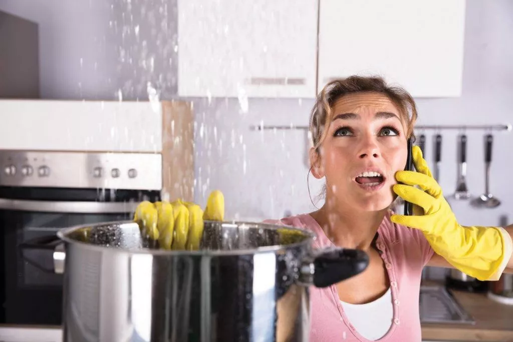 Worried woman talks on phone and holds a large cooking pot to catch water that's leaking from her ceiling