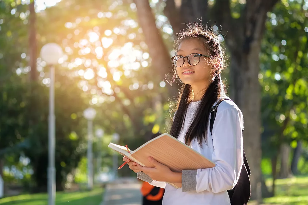 a woman on a college campus looks up, holding her book