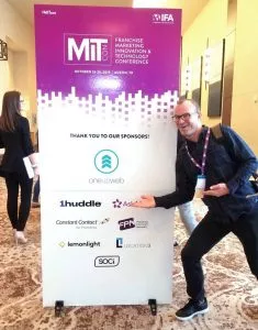 Oneupweb employee at MIT Conference standing in front of poster that contains Oneupweb logo