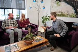 a man and a woman site on a purple couch, listening and taking notes while another man on a purple chair talks to them about a digital marketing budget in an office setting