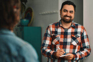 a marketer wearing a checkered shirt leans on a wall and talks to a colleague wile taking notes in a notebook