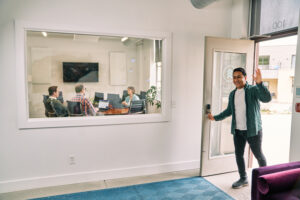 a man walks into a well lit marketing agency office and a meeting is seen through a window in a meeting room